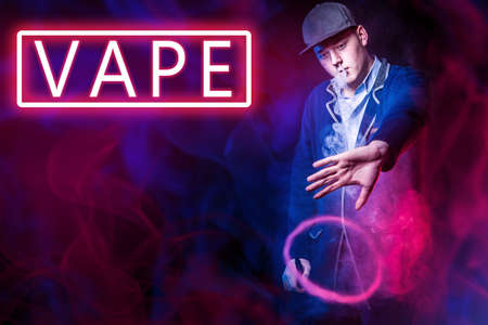 Vape next to man. Smoke cloud in form of a ring. Guy makes a round vape cloud of smoke. Vape neon sign. Steam e-cigarette. Young vaper is in smoke from vaping. Concept - vaping device tricksの写真素材