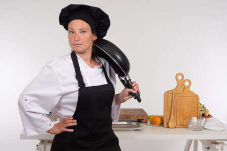 Cook chef woman in restaurant kitchen. Cook is posing with a frying pan in his hands. Chef kitchen. Woman chef on kitchen table background. Concept - career as a cook. Work as a cook in a restaurant.の写真素材