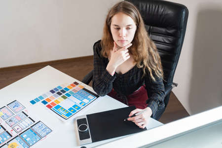 UX designer. Application design. Woman designer uses graphic electronic tablet. Mobile app templates are drawn on paper. Web designer is working on a smartphone app. Girl works in a web design studioの写真素材