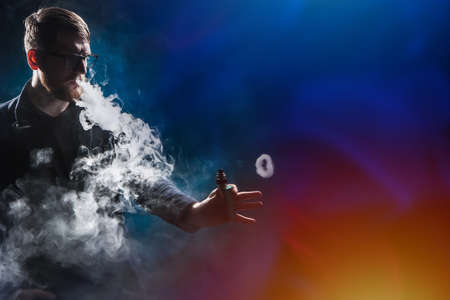 Vaper in puffs of smoke. Man and smoke on a dark background. Ring from an electronic cigarette. Concept - buying a devas for a vaper. The guy is vaping. Components and accessories for vape.の写真素材