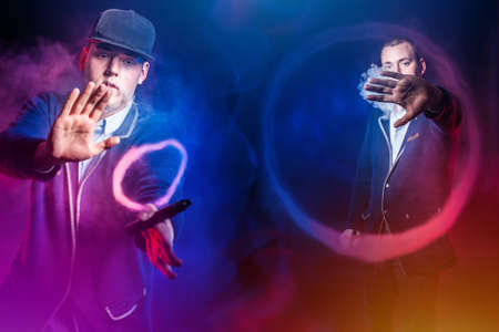 Vape smoke figures. Vapers doing smoke tricks. Two vapers create rings of steam. Concept - chicken vape devices. A man with vape devices on a dark background. Guys push the rings off with their handsの写真素材