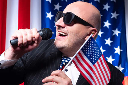 Close-up portrait of American showman. Showman on background of US flags. Man is holding microphone and flag of America. Concept - triotic TV show. Showman covers US voting day or independence day USの写真素材