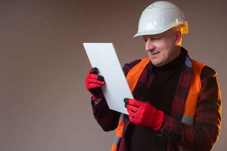 Builder is holding a white tile. Builder is smiling. Man in a building uniform examines something. Concept - sample building material in hands. Concept - installer is satisfied with the qualityの写真素材