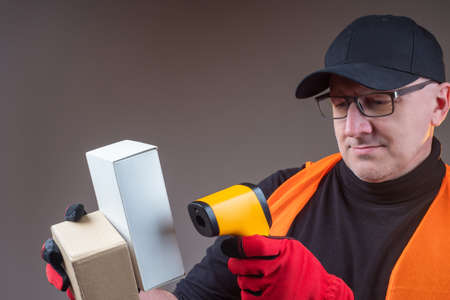 An employee of a postal company. Adult man works with parcels. Concept - a device for reading barcodes. Man registers goods with a scanner. Warehouse worker checks the goods. Man in a capの写真素材