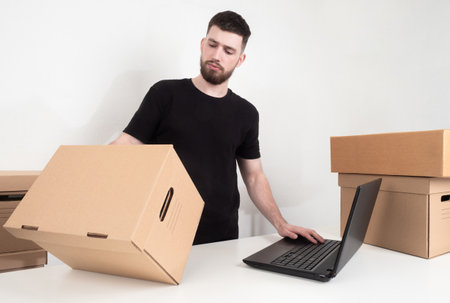 Employee of an online store completes an order. Man holds a box in his hands. Human registers goods using a laptop. Concept - man is preparing goods for dispatch. Guy looks at a cardboard box.の写真素材