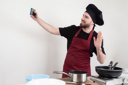 Chef takes a master class on the phone. Cooking training. Cook in a black hat. Cook is making a training video. chef kitchen takes off the recipe. Guy waves his hand. Restaurant chef takes a selfieの写真素材
