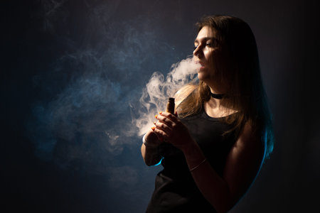 Girl smokes VAPE. Vaping girl. Vaper woman in a cloud of smoke. The girl is holding an electronic cigarette on a dark background. VAPE shop. Vaping concept with space for text.の写真素材