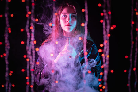 Girl with a VAPE in her hands on the background of garlands. Female Vaper looks at the camera against a dark background. Serious girl with an e-cigarette. The concept of vaping and clouds of smoke.の写真素材