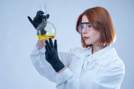Chemist girl. Laboratory assistant examines the substance in the flask. Biochemical research. Analysis of finished products at the enterprise. A woman in a white coat holds a flask in a yellow liquid.の写真素材