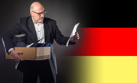 Dismissed man of Germany. Dismissed man holds a box. Unemployed next to flag of Germany. Dismissed man in a business suit. Concept - mass cuts in Germany. German reads a letter of resignationの写真素材