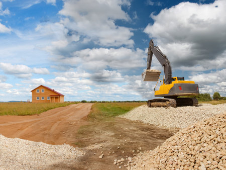 Crawler excavator stands on the stones. Concept - stones unloaded for construction work. Concept - construction of farm. Work as an excavator driver. Demolition services. Wooden house in the distanceの写真素材