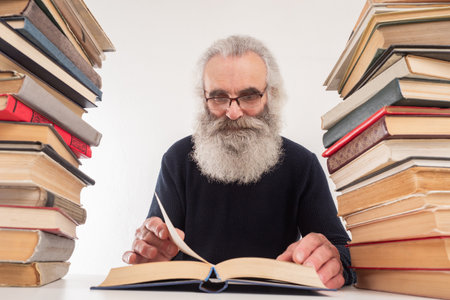 Grandfather is reading books. Grandfather is next to stacks of books. Lot of books on pensioner desk. Concept - never late study. Elderly student. Old man reads a lot. Old man with a gray beardの写真素材