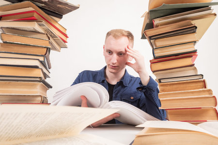 Young guy next to piles of books. Guy is reading something. Young man looking for something in...