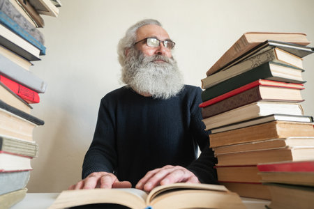 Portrait of old man next to books. Gray-haired old man is reading something. Senior man next to many books. Human is looking for something in books. Senior citizen is studying literature. Wise.の写真素材