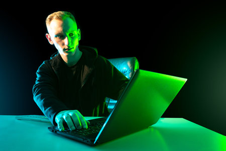 Blogger next to a laptop. Young guy works with a computer. Man and laptop on a dark background. A blogger is writing article. Concept - journalist prints article. Blogger is preparing a publication.の写真素材