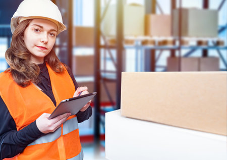 Girl works in a warehouse online store. Delivery of goods from online store. Young girl on background of warehouse. Warehouse worker holds a tablet. Concept - stock accounting application. Auditの写真素材