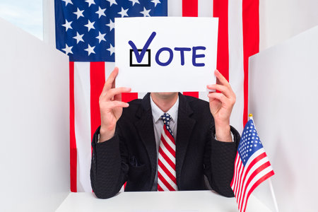 Man in polling station holds VOTE sign. Voter from US shows VOTE logo. man in voting booth. American during USA presidential elections. He calls for participation in elections. US flag in backgroundの写真素材