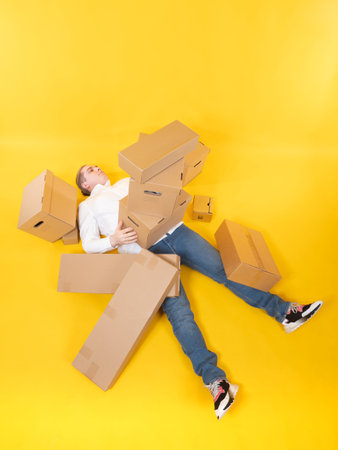 The man is lying on the floor under a pile of cardboard boxes. Metaphor - person is overloaded with...