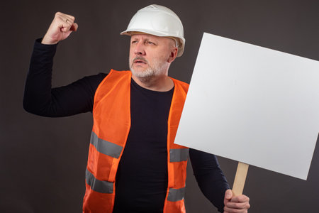 Builder with a banner. Concept - builder shouting slogans. Guy shows a fist. Construction worker in protest. White poster in the hands of a man. Strike in connection with the reduction of employees.の写真素材