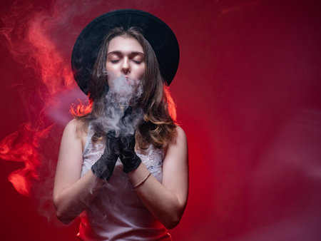 Girl in the hat smokes. Young woman is vaping. Concept - device for vaping. Vaper on a red background. Woman face hides smoke. Human blows steam from vape out of his nose. Girl with long hair smokes.の写真素材