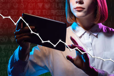Girl follows the stock quotes. Falling stock prices. A woman with a tablet on the background of a falling graph. Situation on the stock market. A woman plays on the stock exchange. Personal finance.の写真素材