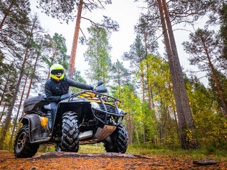 The driver of the ATV is looking at the camera. Man on a ATV in a yellow helmet. A trip through the forest on a ATV. Man is standing next to his qodracycle. Biker pans on the background of the forestの写真素材