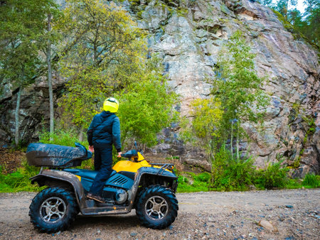 ATV ride. ATV driver looks at the sky. Biker stopped and admires the sky. ATV on the background of the mountain in the forest. Biker admires the top of the mountain. Yellow quadricycle.の写真素材