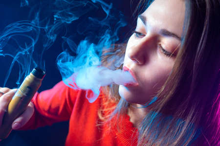 Girl smokes a vape device. Student's face in a cloud of smoke. Woman's face with a cigarette close-up. Electronic cigarette in the hands of a girl. To let out a mouth from lips. Store for the vaperの写真素材
