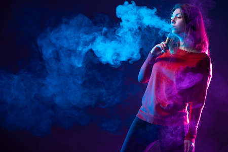 Woman viper on a dark background. Vaper in the clouds of steam. Steam next to a smoking woman. Concept - modern cigarettes. Concept - giving up cigarettes in favor of vape. Young girl with an e-cigの写真素材