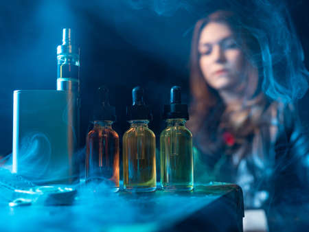Vape devices are on the table. The oil next to the electronic cigarette. Bottle vaping liquids. A viper girl in the background. Woman in the smoke from vaping. Concept - vape oils of different tastesの写真素材