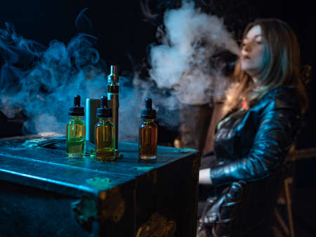 Devices for vaping. Vape oils are on the table. Vape liquids next to the vaper. A device for smoking next to bottles of juice. Concept - a store for vapers. Girl vapit in the backgroundの写真素材