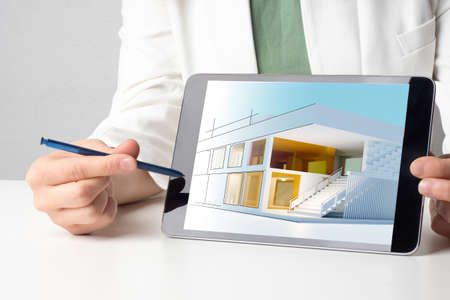 Building on tablet screen. Architectural project. New cottage layout. A person demonstrates a mock-up of a house on a tablet computer. A construction engineer shows a drawing of a house on a tablet.の写真素材