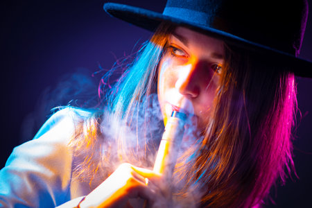 Girl smokes a vape and looks away. Concept - youth cigarettes. Vaper blows steam through his nose. Woman vaper lets off steam and nose. Female student in a hat with an electronic cigaretteの写真素材