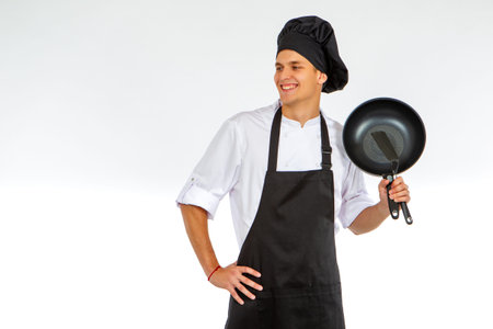 Young cook is holding a frying pan. Chef guy on a white background. Man in a cook's uniform smiles....