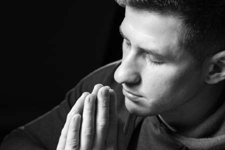 Close-up of a praying man face. Young man praying to god. Catholic or Christian during prayer. Praying guy on a black background. He fold his palms to read prayer. Catholic asks god for helpの写真素材