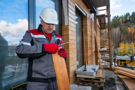 Builder is engaged in exterior building. He works on wood. Builder prepares wood house cladding. Concept finishing house with wood siding. Male builder during work. House facade cladding with sidingの写真素材