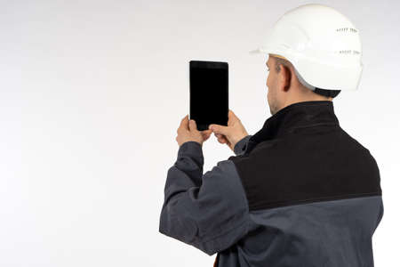 Worker takes photographs of something on phone. Man photographs something with a smartphone. Concept - he makes a photo report for the employer. Worker in a gray uniform. Man in a white safety helmetの写真素材