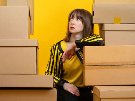 Business woman is thinking about something. Portrait in middle of boxes. She looks up thoughtfully. Business owner woman. Girl and boxes on a yellow background. Concept - she is waiting for courier.の写真素材