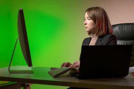 Businesswoman works in office. She uses a computer and a laptop same time. Businesswoman at office table. Woman is typing something. Office worker on green background. Concept - businesswoman careerの写真素材