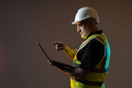 Worker stands and looks at the information in the laptop. An engineer in a work uniform and a white hard hat. A man checks his e-mail on a laptop. The architect brings his finger to the touch screen.の写真素材