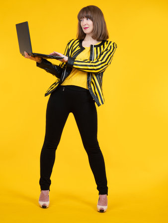 Cheerful young woman. She isholding laptop in her hands. Happy girl with laptop on yellow background. Happy freelancer girl with laptop. Brown-haired woman is dressed in casual style. Studio portraitの写真素材