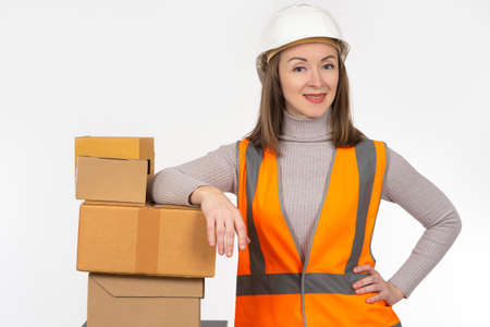 Female storekeeper leans against a stack of cardboard boxes and smiles at the camera. Girl in a white helmet and a reflective vest. Working in a warehouse. Warehouse accounting. Home delivery of goodsの写真素材