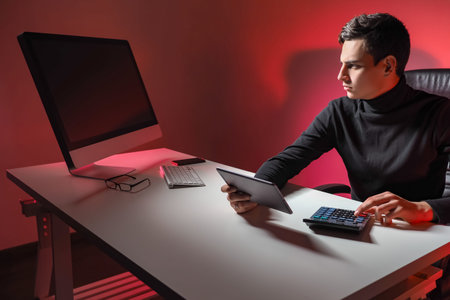 Accountant looks at off monitor. He is holding a tablet computer and calculator. Young guy in...