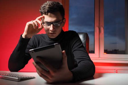 Young investor with a tablet computer. He invests through a tablet app. Guy investor with glasses sits at table. Application for Investing in stock exchange. Investing in stocks over Internet.の写真素材