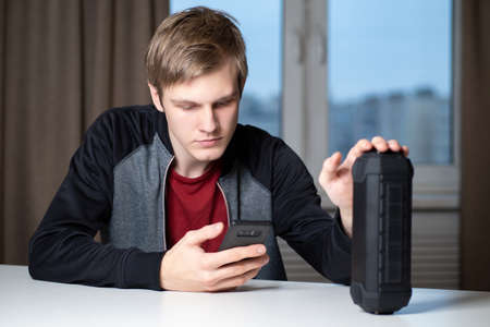 Young guy turns on the speaker. He controls the portable speaker through his smartphone. Portrait of a student with a portable column. Concept - intelligent voice-activated speaker.の写真素材