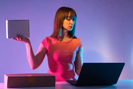 Girl with a box in her hands. She works in her owner business. Girl owner of a business on internet. Concept - woman sells goods on Internet. Businesswoman with laptop and boxes on lilac backgroundの写真素材