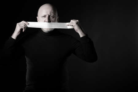 Man covers his mouth with duct tape. Concept he doesn want to let it slip. Human with scotch tape on black background. Man is trying to keep secret. He is afraid to say something. Tries to be silentの写真素材