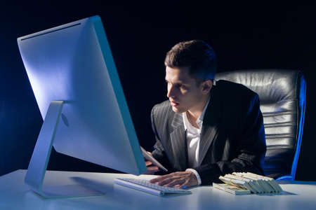 Man looks aggressively at computer screen. Bundles of money next to a businessman. Concept - he works as a trader. Trader works at an office desk. Trader in a dark office. Earned money by his sideの写真素材