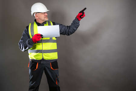 Builder on a dark background. Man in a builder uniform looks up. He points his finger to the side. Builder work concept. Career in a construction company. Worker holds a sheet of paper in his handsの写真素材