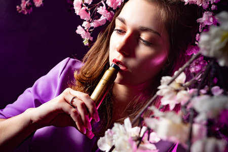 Student is going to smoke a vape. Girl brings a vape device to her mouth. Vaper. Concept - eco friendly cigarettes. Eco friendly alternative to smoking. Viper girl next to the flowers. Blooming treeの写真素材